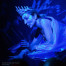 A performer wearing a jeweled crown and elaborate blue makeup leans over a miniature ship model, bathed in blue light.